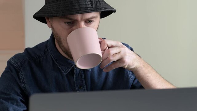 Man at desk lifts pale pink mug then refocuses on laptop in intimate workfromhome closeup, with man, mug, sip, coffee detail, showing cup, laptop, desk, hat context