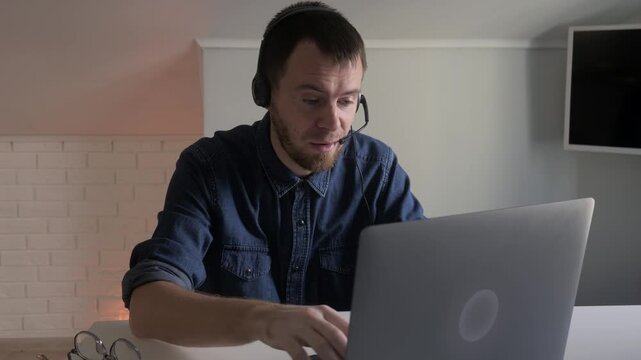 Young man wearing headset typing and speaking into laptop during remote work call in home office with brick accent wall and warm side light, with man, headset, microphone, laptop detail
