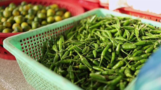 Close-up of fresh green chili peppers and citrus fruits displayed in plastic baskets on a stall at Xom Moi Market in Nha Trang, Vietnam. Commercial food supply and retail trade concept.