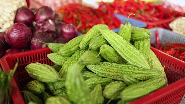Close-up of fresh bitter melons, red onions and chili peppers displayed in plastic baskets on a stall at Xom Moi Market in Nha Trang, Vietnam. Commercial food supply and retail trade concept.