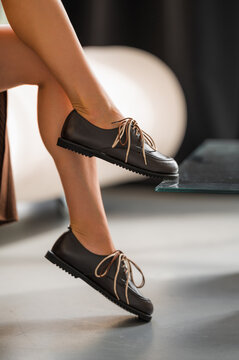 Closeup of crossed female legs wearing brown leather laceup shoes with contrast laces, resting beside glass table edge in softly lit modern interior with blurred background and elegant fashion mood