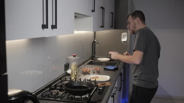 Man handling eggs beside gas cooktop in modern kitchen with cast iron skillet, open carton, oil bottle, white bowl, reflective backsplash, and focused preparation for cooking
