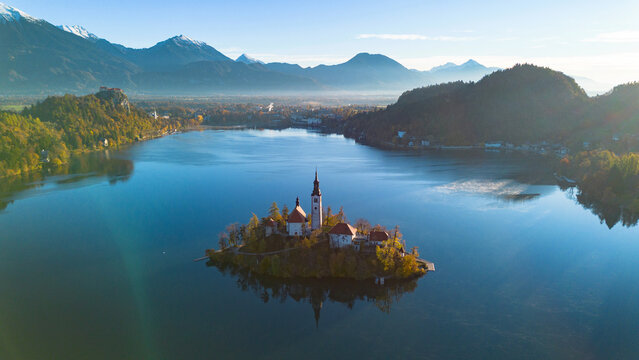 Aerial view of Bled Island and the Church of the Assumption rising above the calm waters of Lake Bled, surrounded by forested hills and the Julian Alps in Slovenia.