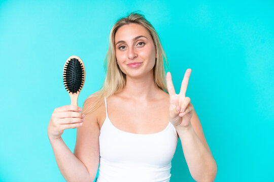 Young caucasian woman with hair comb isolated on blue background smiling and showing victory sign