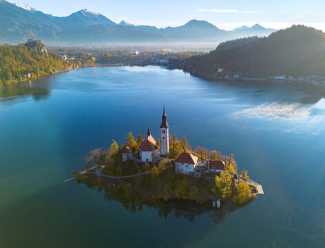 Aerial view of Bled Island and the Church of the Assumption rising above the calm waters of Lake Bled, surrounded by forested hills and the Julian Alps in Slovenia.