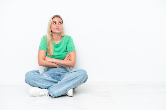 Young caucasian woman sitting on the floor isolated on white background making doubts gesture while lifting the shoulders