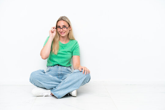 Young caucasian woman sitting on the floor isolated on white background with glasses and happy