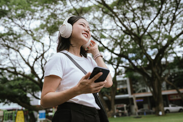 Asian woman wearing wireless headphones walking outdoors while using a smartphone, representing modern lifestyle, mobile technology, music, communication, and urban leisure. © Kieng
