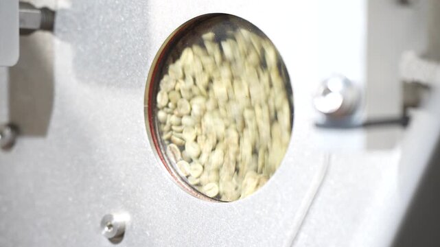 Green coffee beans churn behind a round viewing port on the roaster's housing, agitating just ahead of the roasting cycle starting.
