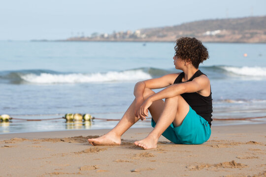 A teenager is sitting on a sandy beach by the ocean, looking at the sea and enjoying a peaceful summer day. The concept of leisure, travel, relaxation, freedom, mindfulness, solitude