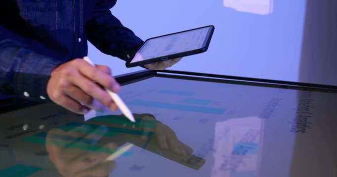Business professional uses a stylus on an interactive touchscreen table while holding a smartphone, viewing colorful charts in a modern blue-lit office.