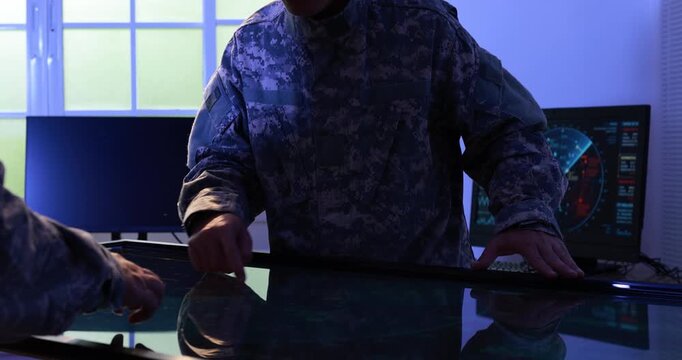Military personnel use a large touchscreen table in a dark command center, with digital maps and radar screens glowing in the background.