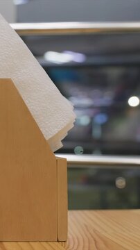 Close up of wooden paper towel holder with textured white tissue. Background is softly blurred showing indistinct shapes. Focus remains on smooth wood grain texture in sharp detail