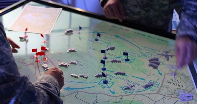 Military personnel plan a tactical operation around an illuminated map table with miniature vehicles, aircraft and red and blue flag markers.