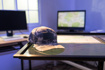 Naklejka premium Digital camouflage military cap resting on a map table in a command office, with computer monitors in the background.