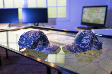Naklejka premium Two digital camouflage military caps rest on a glass strategy table with maps, computer monitors and a wall display in a modern command center.