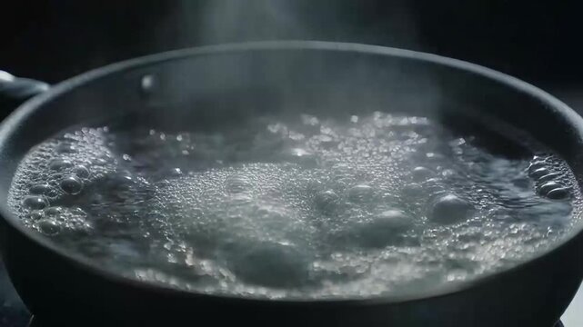 Closeup video of water boiling rapidly in a dark matte cooking pan, the bubbles roll and collide like living organisms under brilliant studio lights