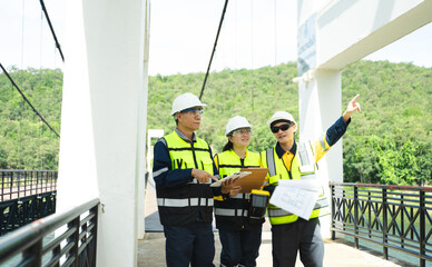 Professional Asian civil engineers in safety vests and hardhats holding blueprints and pointing up...