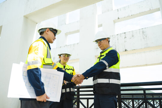 Asian civil engineers shaking hands in agreement after reviewing blueprint plans on an outdoor suspension bridge construction project.