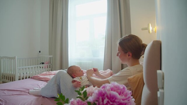Careful maternal touch opens infant clenched fist while baby rests on bed. Soft finger movement encourages hand development, tactile awareness, coordination, trust, and attentive home caregiving.
