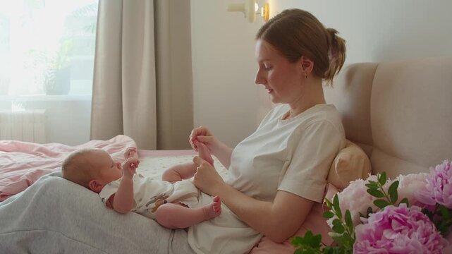 Mother talks softly while massaging baby toes during calm time on bed. Gentle touch and warm voice encourage relaxation, sensory awareness, emotional connection, and patient home caregiving.