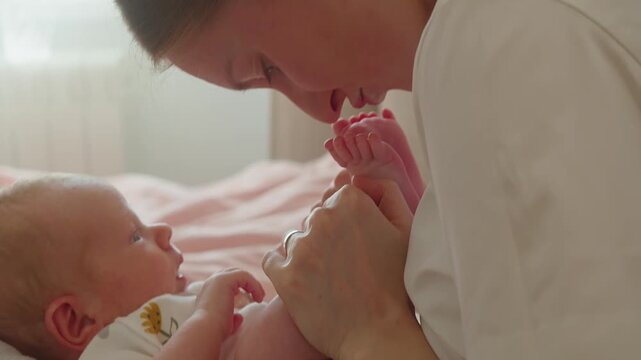 Mother gently kisses infant foot during affectionate play on bed at home. Tender gesture expresses love, warmth, comfort, trust, emotional closeness, and caring maternal connection.