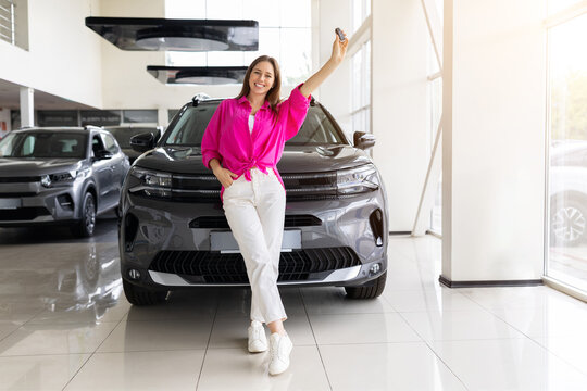 Smiling young woman poses in front of new SUV at dealership showroom while raising the car key, expressing excitement, confidence, and satisfaction with her purchase