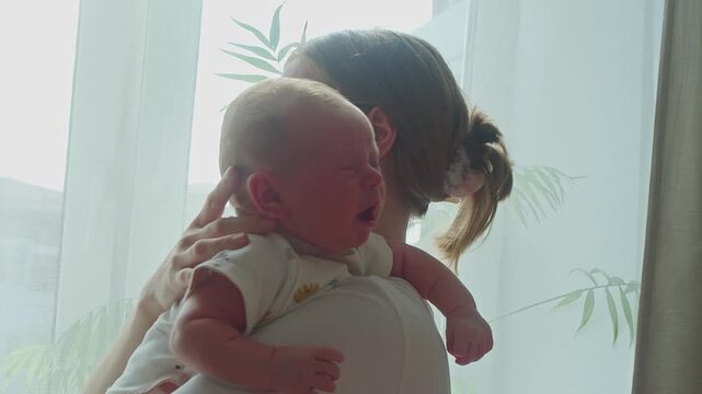 Mother approaches window with crying baby, seeking gentle mood lift from sunlight while keeping infant close. Natural brightness may ease distress and support patient maternal soothing.