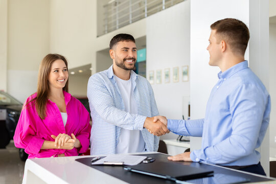 Salesman shaking hands with smiling young couple at showroom, finalizing car purchase deal at dealership counter, celebrating a successful agreement and completed vehicle buy