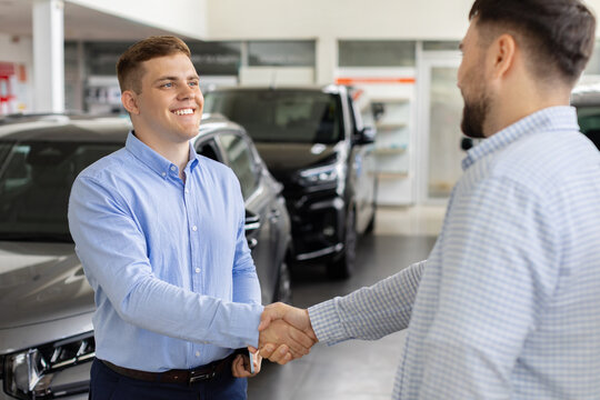 Smiling car sales consultant shaking hands with young male customer in dealership showroom after reaching an agreement and successfully completing a vehicle purchase