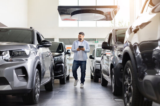 Young male customer walking through modern car dealership with tablet, comparing displayed vehicles and researching models, features, and options before buying
