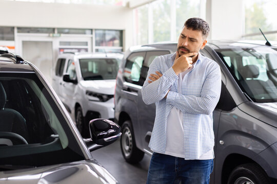 Thoughtful young male buyer examining car in dealership showroom, considering vehicle design, features, model options, and preferences before making a purchase decision