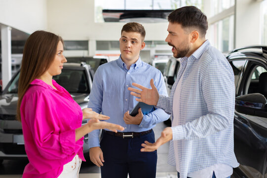 Confused salesman listening as young couple arguing and gesturing emotionally during car negotiation at dealership showroom