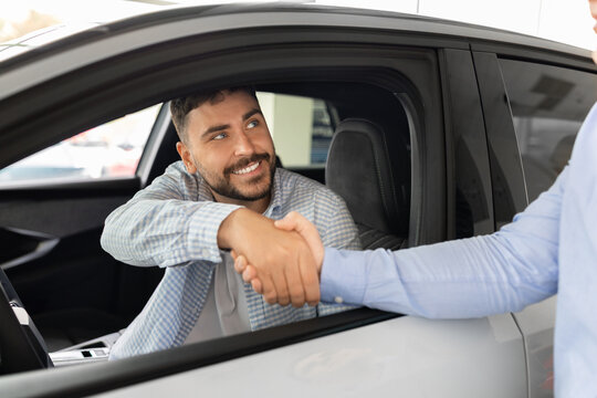 Young male buyer shaking hands with car sales consultant through the open window of a showroom vehicle, confirming a successful deal, purchase agreement, and customer trust