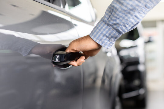 Close-up of young male customer opening car door in dealership showroom, inspecting the vehicle exterior, handle design, accessibility, and functionality