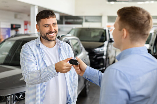 Happy young male buyer receiving car keys from sales consultant inside modern dealership showroom, marking vehicle delivery, ownership, and successful purchase