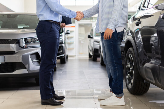 Car salesman and young male buyer shaking hands beside vehicle in dealership showroom, symbolizing agreement, successful negotiation, purchase, and customer trust