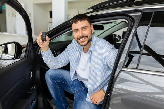 Smiling young male buyer sitting inside new car at dealership and proudly shows the vehicle key, celebrating successful purchase, ownership, delivery, and driving freedom