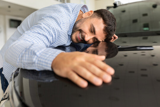 Happy young male buyer affectionately leaning on new car in dealership showroom, expressing excitement, satisfaction, and emotional attachment after choosing his vehicle