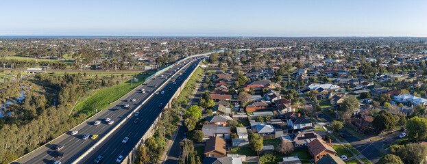 Obraz premium Aerial&nbsp;panorama view of Princes Freeway traffic bordering suburban residential housing, green reserves, and a railway line in Laverton, Melbourne, Australia. Traffic parallel to rail infrastructure