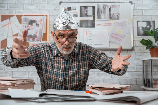 Agitated older man wearing an aluminum foil hat explaining a conspiracy with an intense expression and evidence boards behind him