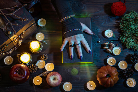 Witch's hands witchcrafting with a wizard's black book with a green glow, casts a spell among candles, pumpkins on Halloween night