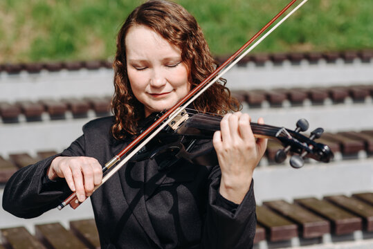violinist outdoors, young beautiful redhead caucasian woman in jacket playing black violin in city park with green grass and trees in sunny summer day, classic music concept