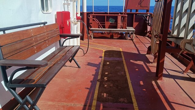 Benches and equipment are arranged on an open cargo ship deck beneath bright daylight at sea. The image represents everyday shipboard life, maritime work, rest, routine, and functional industrial
