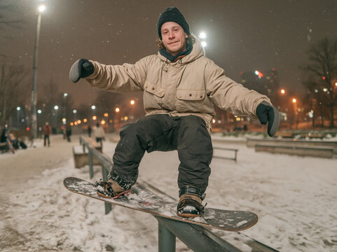 Young male snowboarder grinding on rail in urban snow park during nighttime snowboarding session