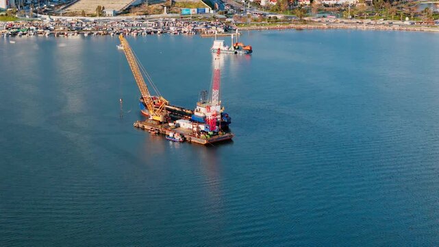 Floating crane barge and dredging vessel working in coastal water near urban waterfront. Aerial view of seabed sediment removal and marine engineering operation