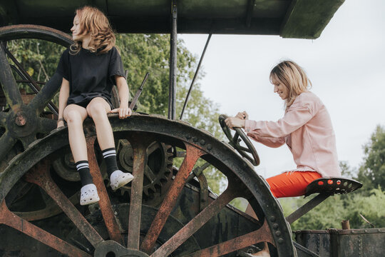 Young girl and woman seated on vintage tractor, with large wheels and green foliage in the background, showcasing a playful outdoor scene