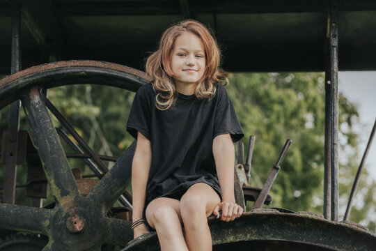 Young girl with wavy hair wearing a black t-shirt sits on a vintage tractor wheel in a green outdoor setting with trees in the background