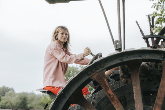 Female operator in pink jacket drives vintage tractor with large wheels in a rural landscape, green trees and cloudy sky visible in the background