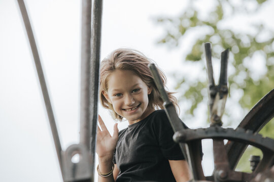 Young girl with brown hair wearing a black shirt smiles and waves while sitting on a vintage machinery wheel outdoors with greenery in the background
